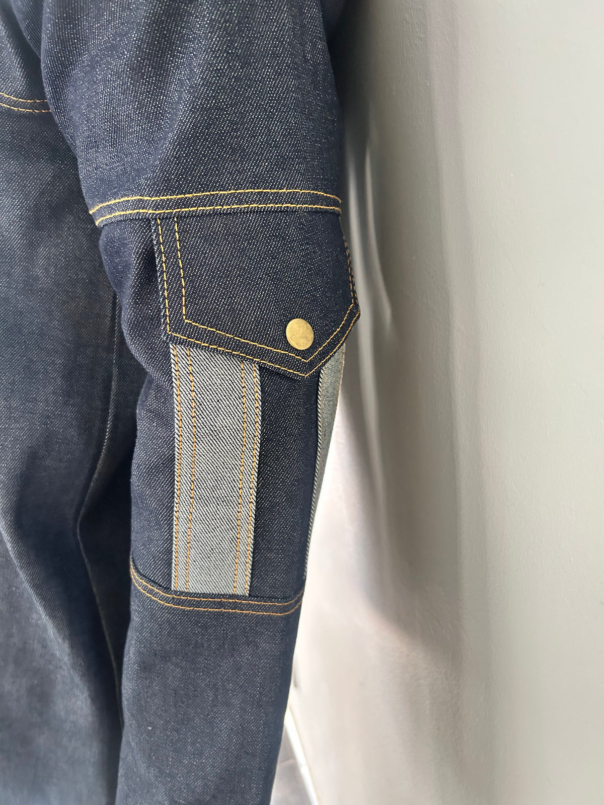 Side close-up of a dark blue denim jacket on a mannequin, showing a grey shoulder stripe, central pleat and contrast stitching.