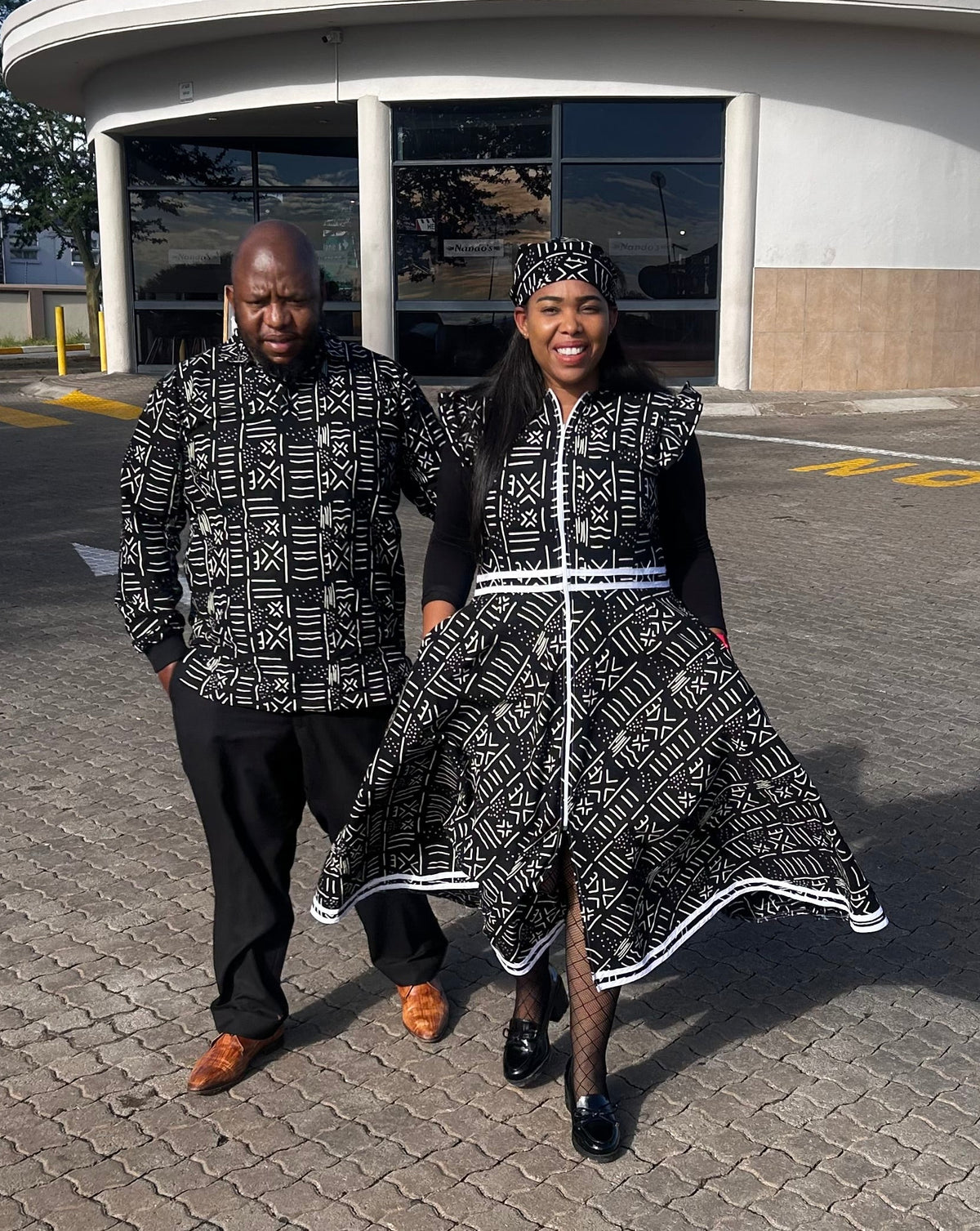 A man and a woman wearing matching black and white patterned traditional clothing walk together in a paved parking lot in front of a building with a curved facade and large windows.