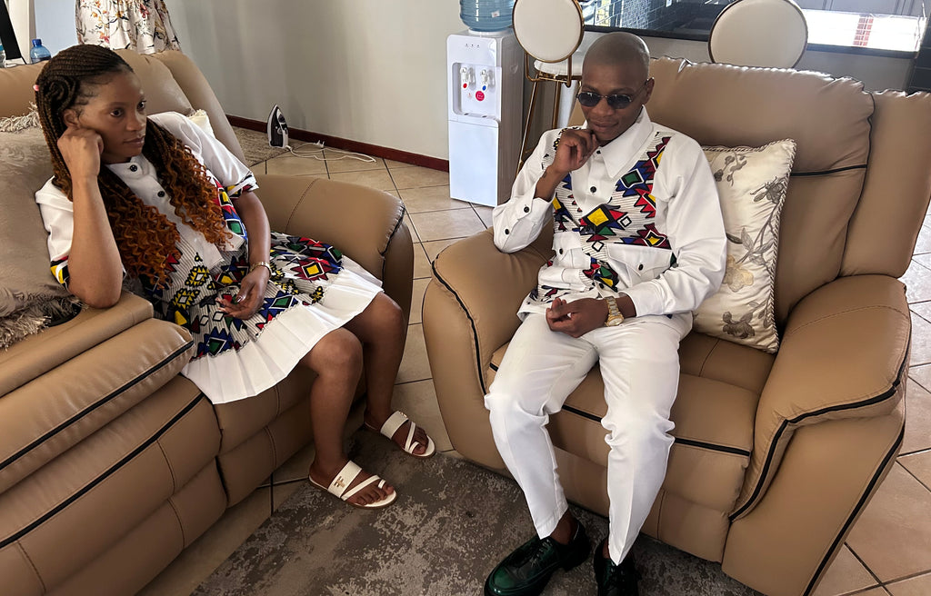 Seated view of a woman in a white top and pleated skirt with colourful Ndebele patterns, beside a man in a matching suit.
