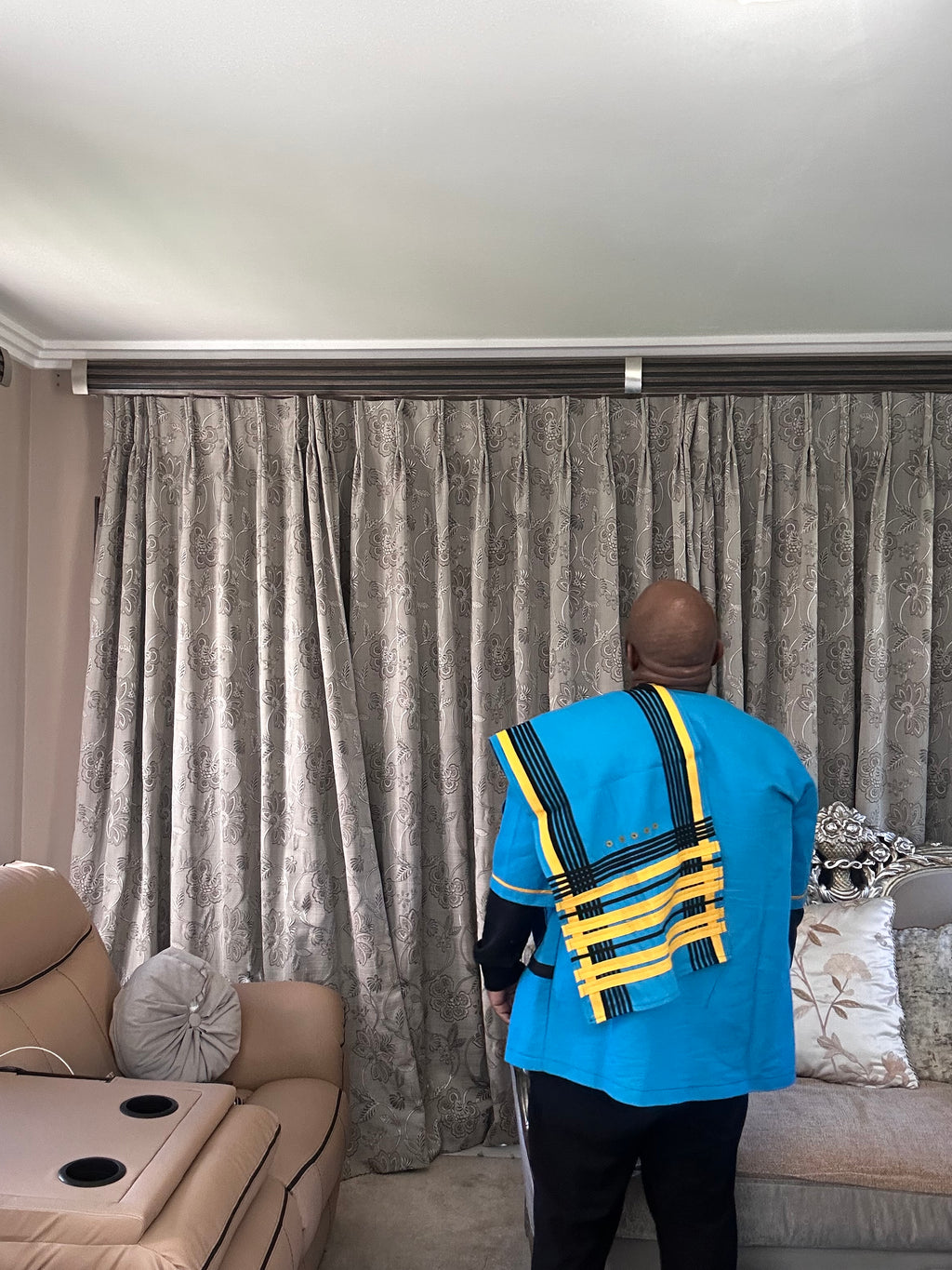 A man viewed from behind wearing a bright blue Xhosa traditional robe with yellow and black woven patterns, standing in front of a large gray patterned curtain in a living room.