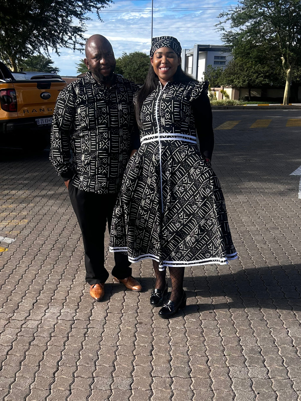 A man and a woman standing together on a paved outdoor street wearing matching black and white patterned traditional clothing, with a yellow pickup truck and trees in the background.