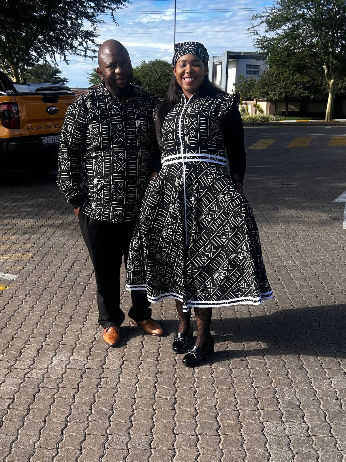 A man and a woman standing together on a paved outdoor street wearing matching black and white patterned traditional clothing, with a yellow pickup truck and trees in the background.