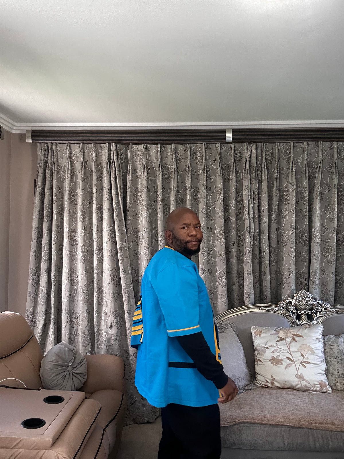 A man wearing a bright blue and black traditional-style Xhosa robe with yellow accents stands in a living room in front of gray patterned curtains.