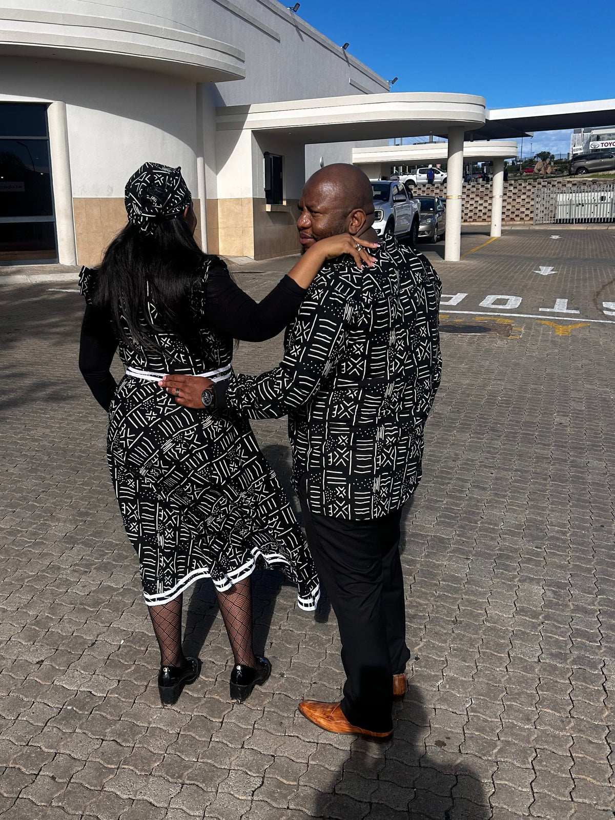 A man and a woman wearing matching black and white patterned outfits pose together in a paved parking lot in front of a white commercial building under a clear blue sky.