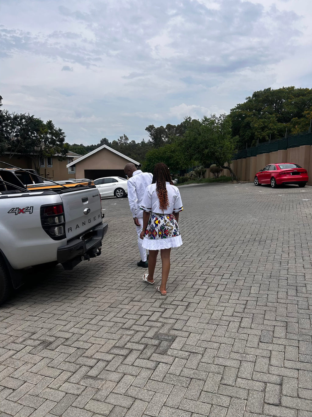 Two people walking on a driveway wearing matching Ndebele patterns with vehicles and trees in the background