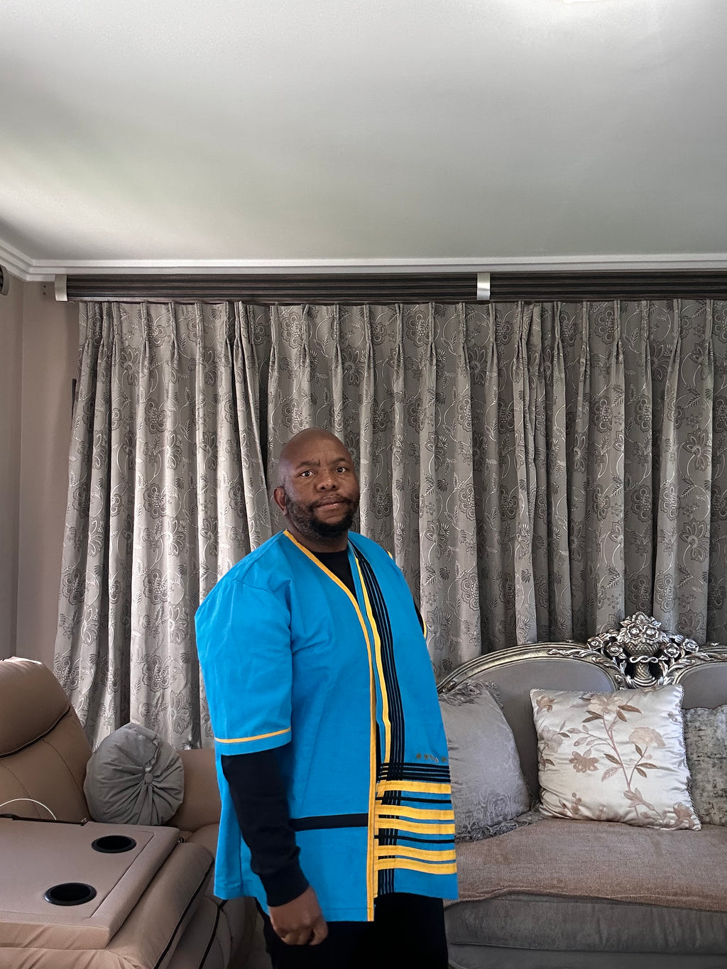 A man wearing a bright blue traditional-style Xhosa top with yellow and black trim, standing in a living room in front of gray patterned curtains.