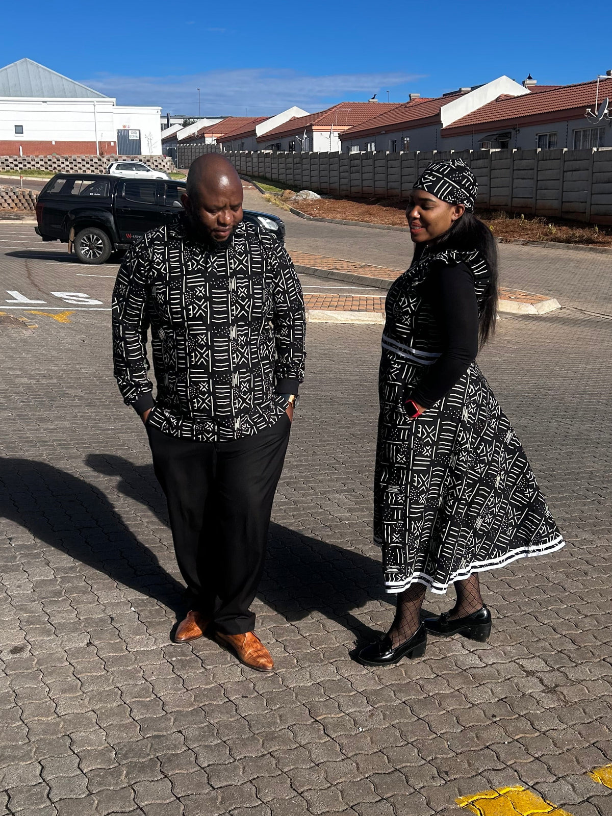Side view of a woman in a black geometric-print dress with a flared skirt and matching headwrap, beside a man in a matching shirt.
