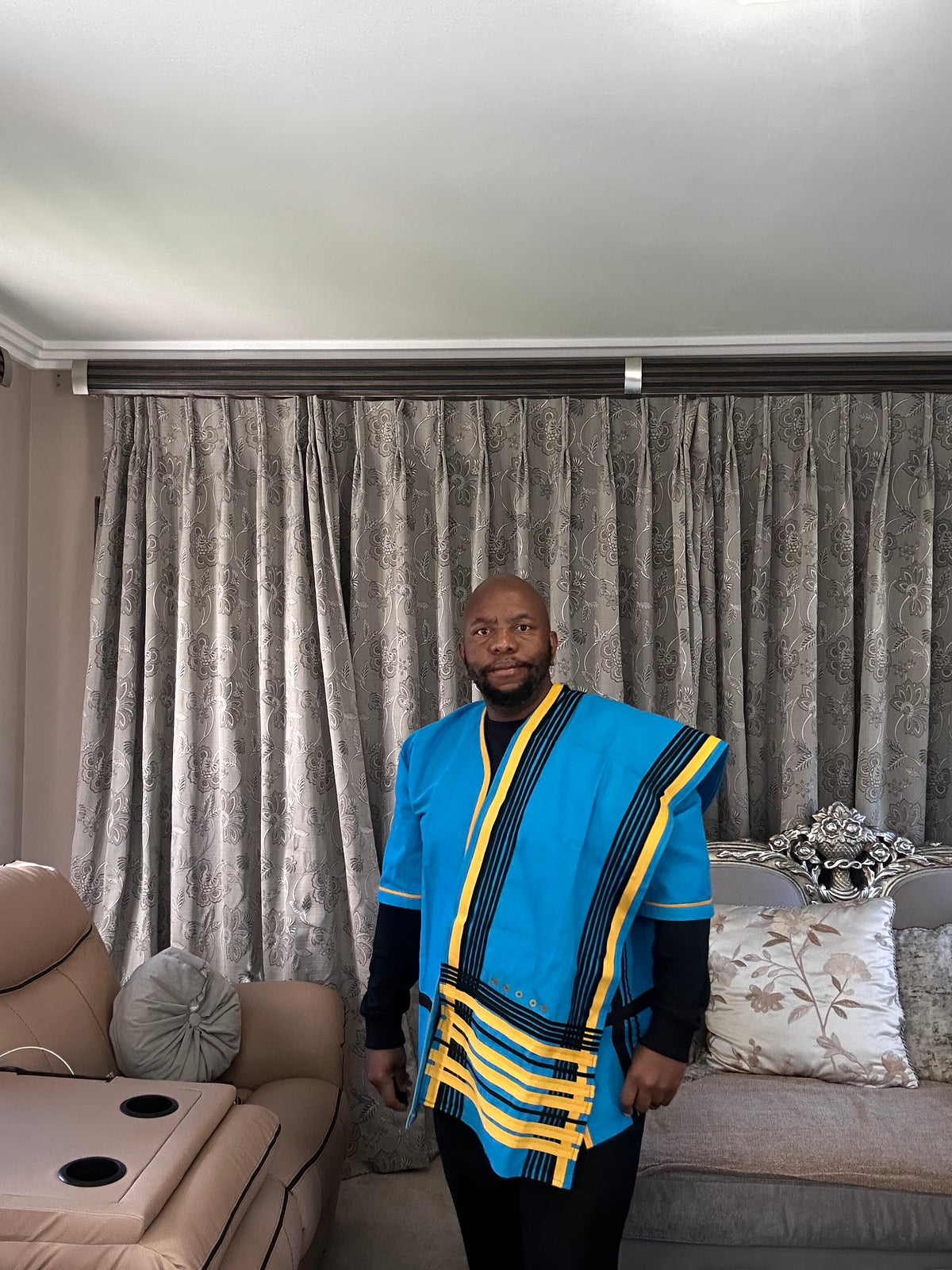 A man wearing a vibrant blue traditional African shirt with yellow and black patterned borders, standing in a living room in front of gray patterned curtains.