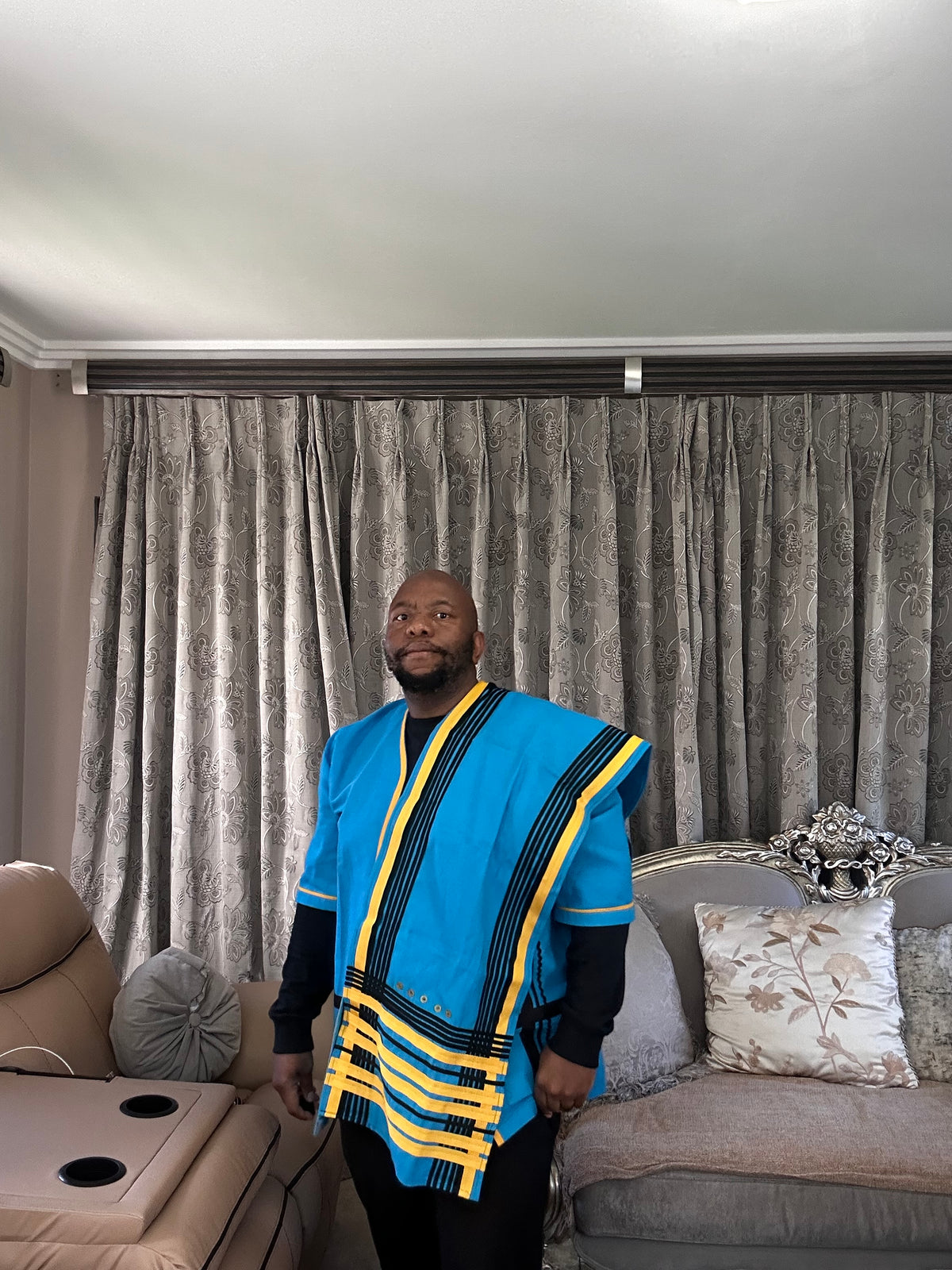 A man wearing a vibrant blue African dashiki with yellow and black stripes, standing in a living room in front of gray patterned curtains.