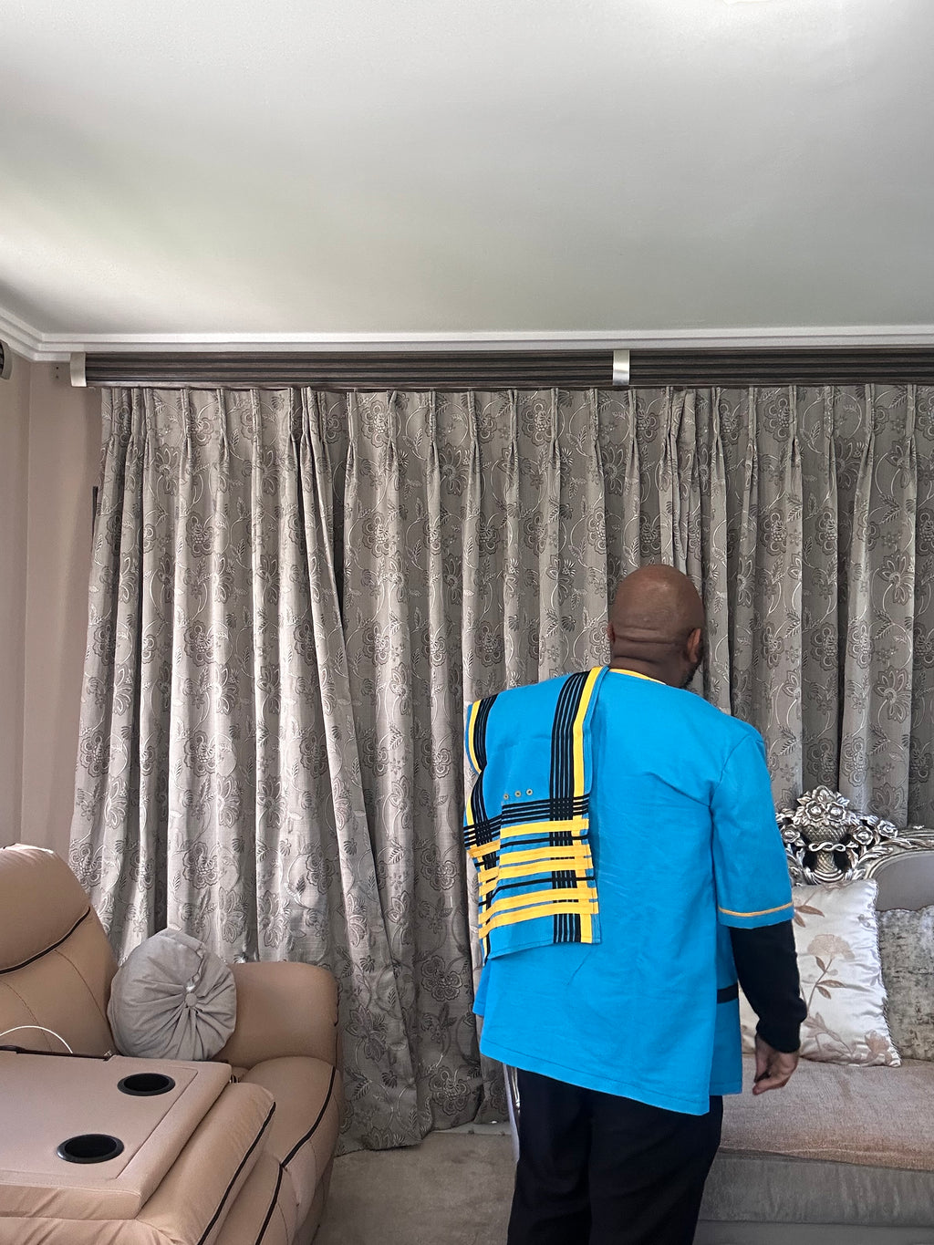 A man viewed from behind wearing a bright blue Xhosa shirt with yellow and black striped shoulder accents, standing in front of a large gray patterned curtain in a living room setting.
