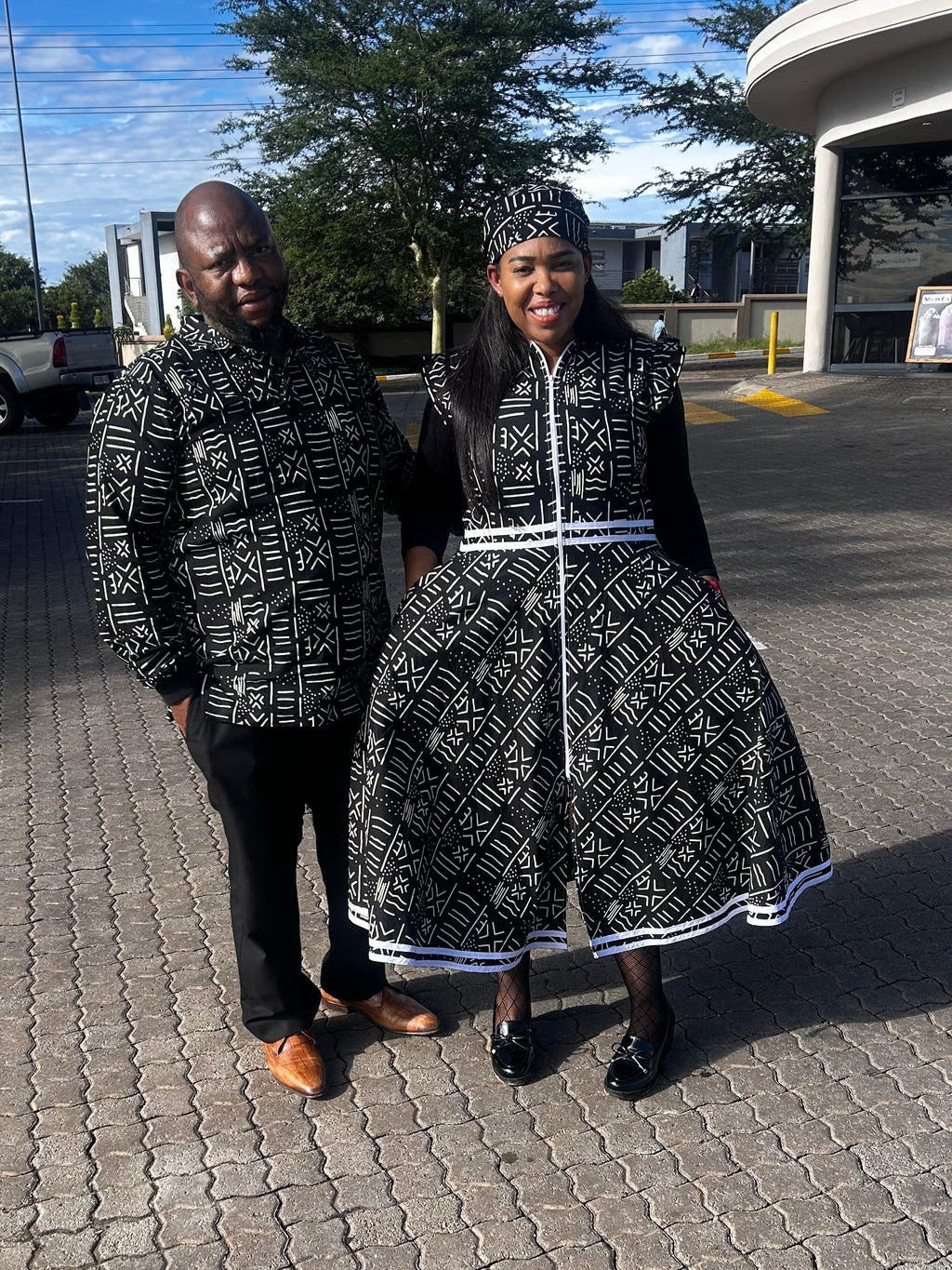 A man and a woman standing together in a paved outdoor parking lot. Both are wearing matching black and white patterned traditional-style outfits with head coverings.