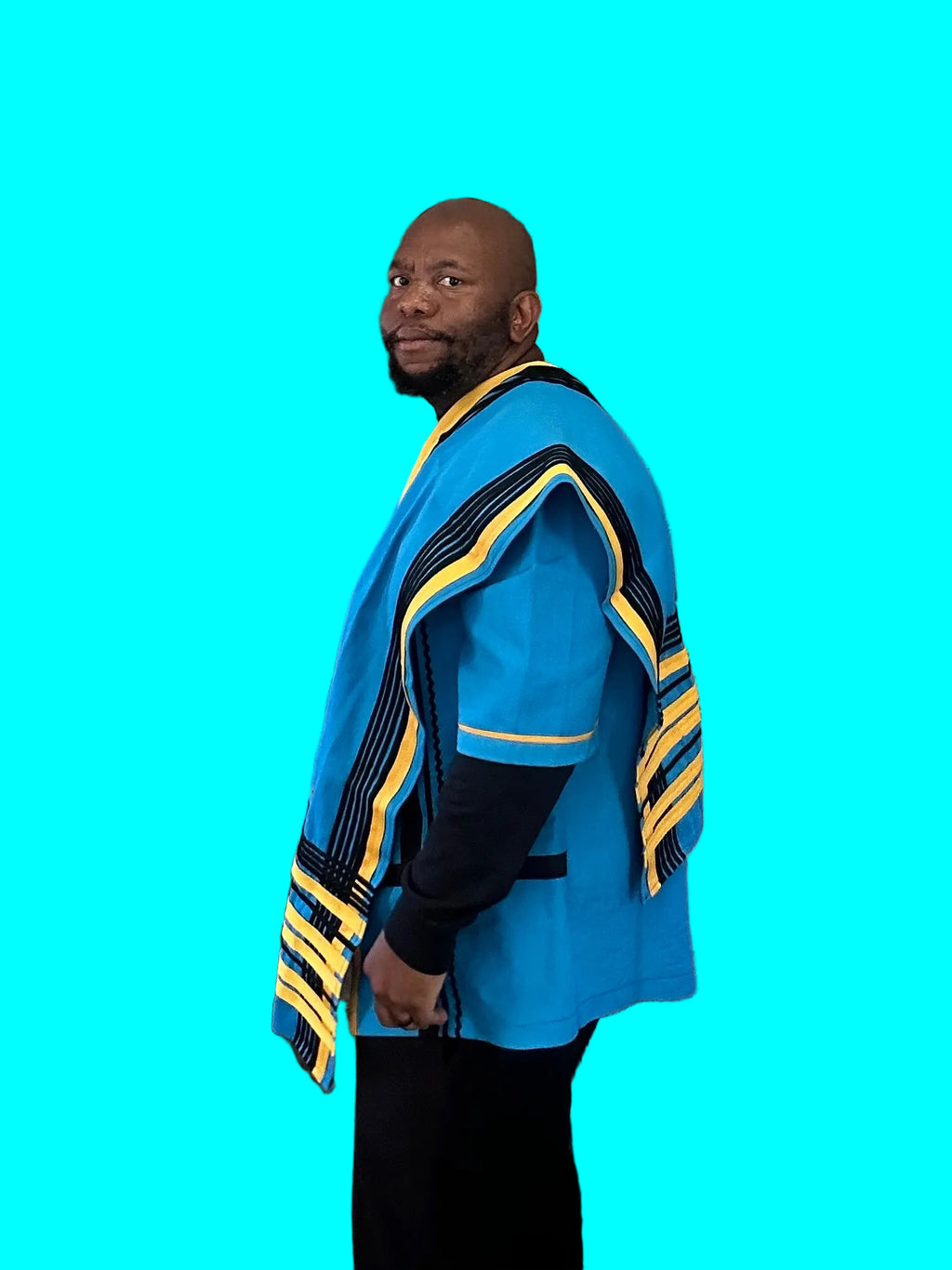 Side view of a man wearing a blue Xhosa top and draped shawl with black and yellow striped trim, styled with black trousers.