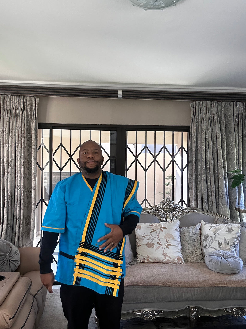 A man wearing a vibrant blue traditional African-style tunic with yellow and black patterned trim, standing in a living room in front of a sofa and window.