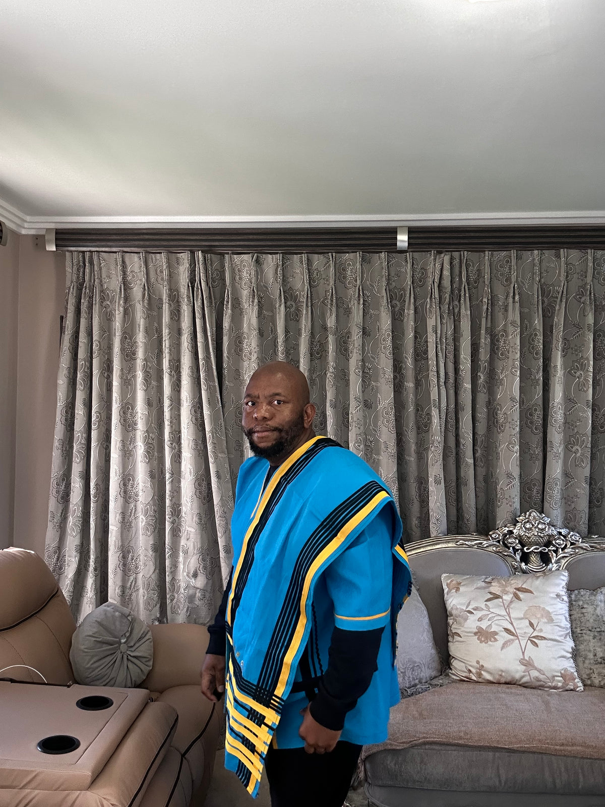 A man wearing a vibrant blue shawl with yellow and black striped borders stands in a living room in front of gray patterned curtains.