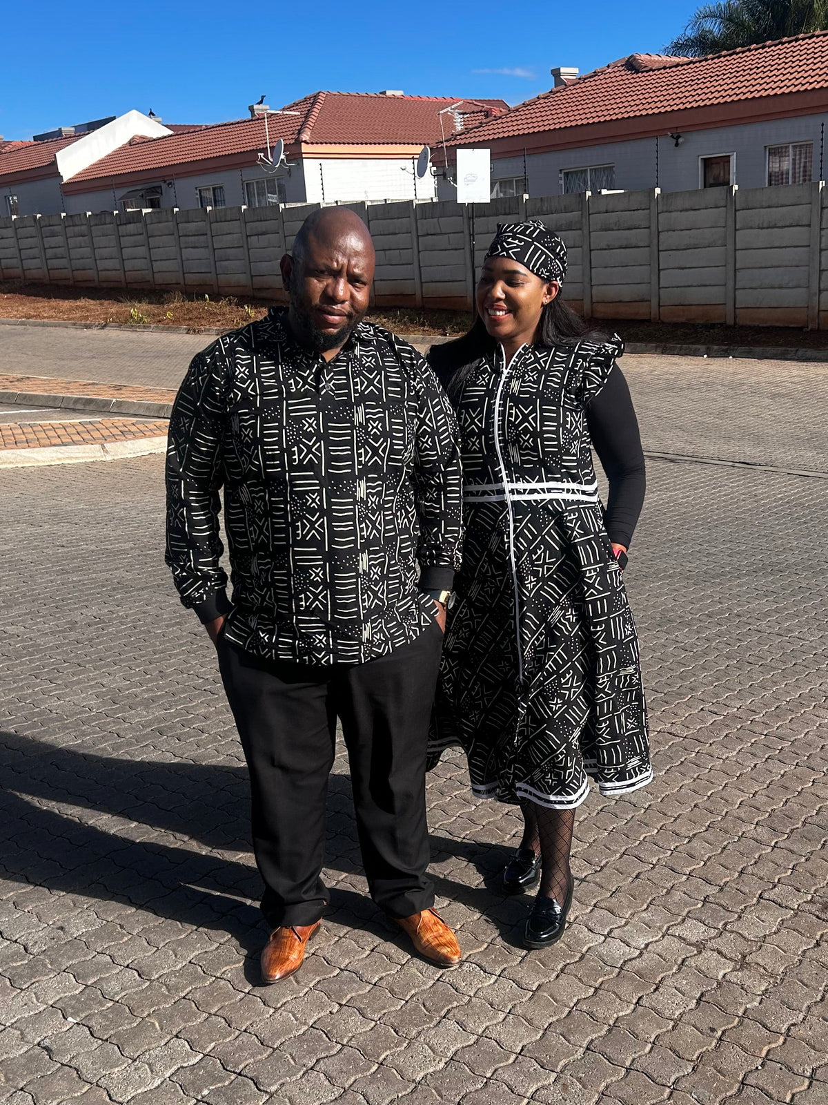 A man and a woman standing outdoors on a paved street wearing matching black and white patterned traditional clothing under a clear blue sky.