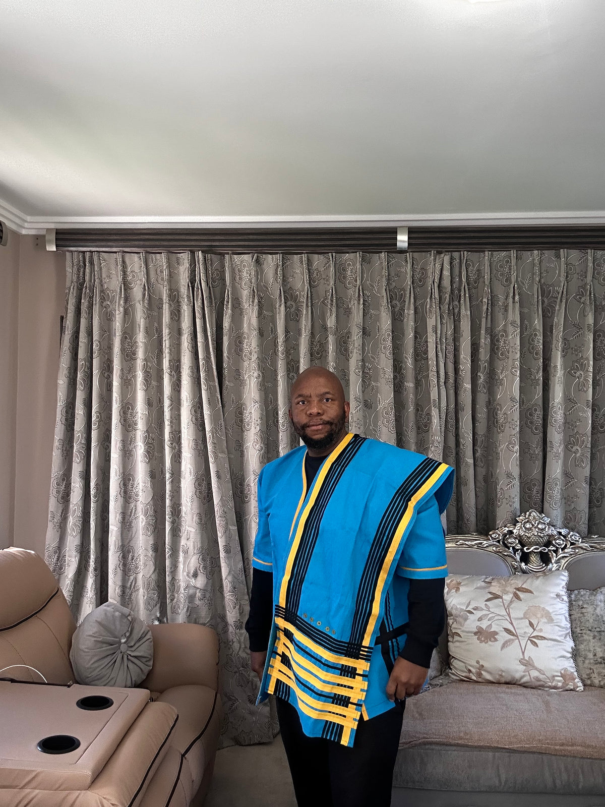 A man wearing a vibrant blue African Xhosa dashiki with yellow and black stripes, standing in a living room in front of gray patterned curtains.