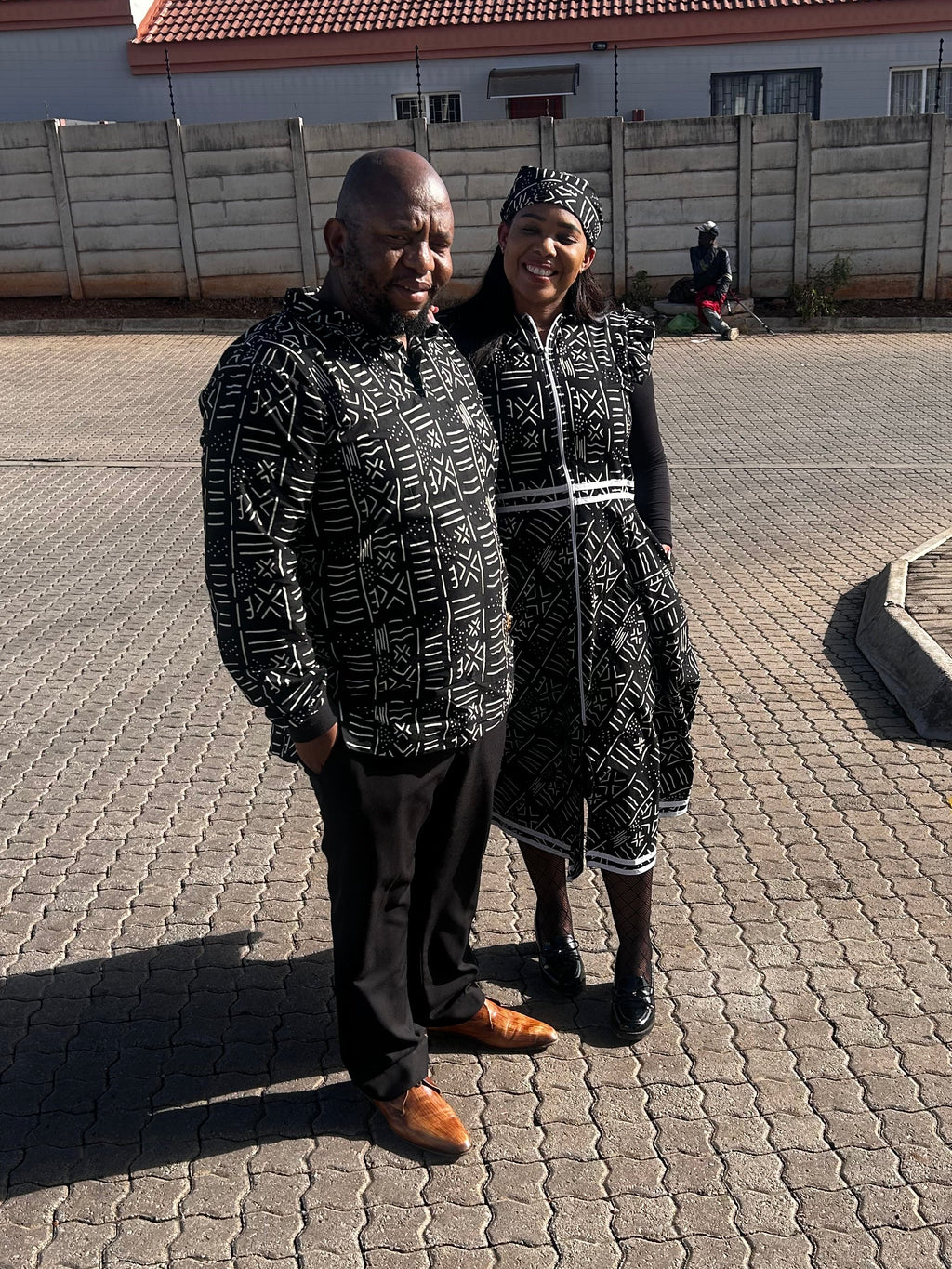 A man and a woman standing together outdoors on a paved street in front of a building with a red-tiled roof and a concrete wall. Both are wearing matching black and white patterned traditional-style outfits.