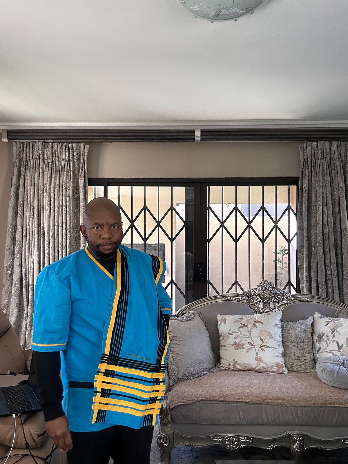 A man wearing a vibrant blue traditional-style jacket with yellow and black striped accents, standing in a living room with a gray sofa and curtains behind him.