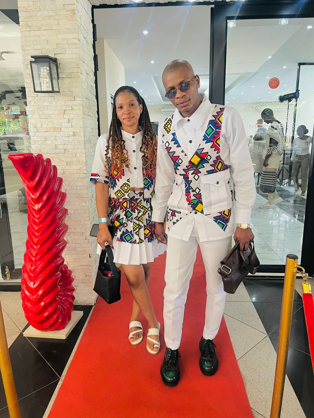 Woman in a white cropped top and pleated skirt with colourful Ndebele patterns, holding hands with a man in a matching suit.