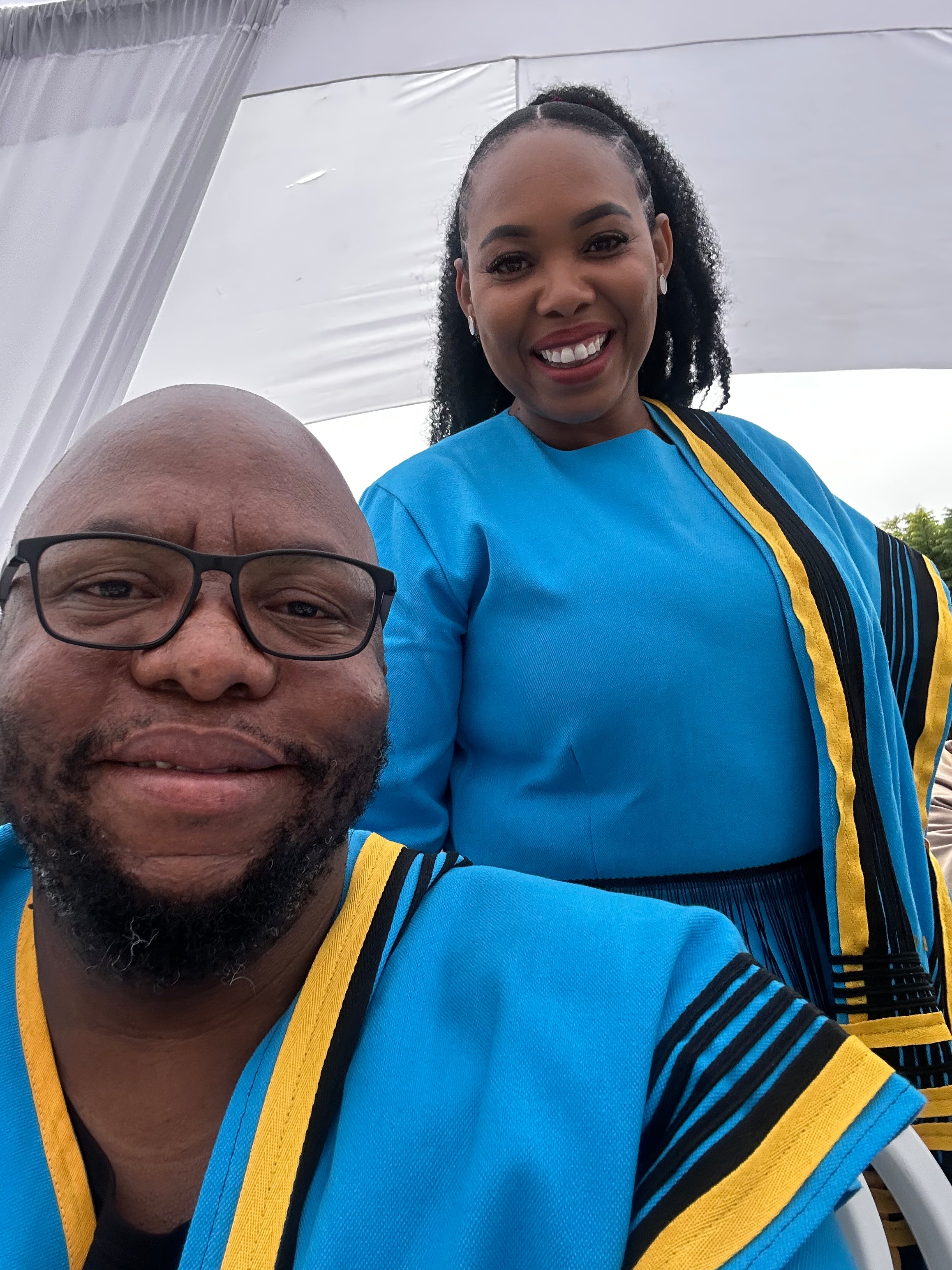 A couple wearing a matching Xhosa outfit in blue gowns with yellow accents standing under a white tent.